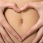 womans hand forming a heart symbol around navel